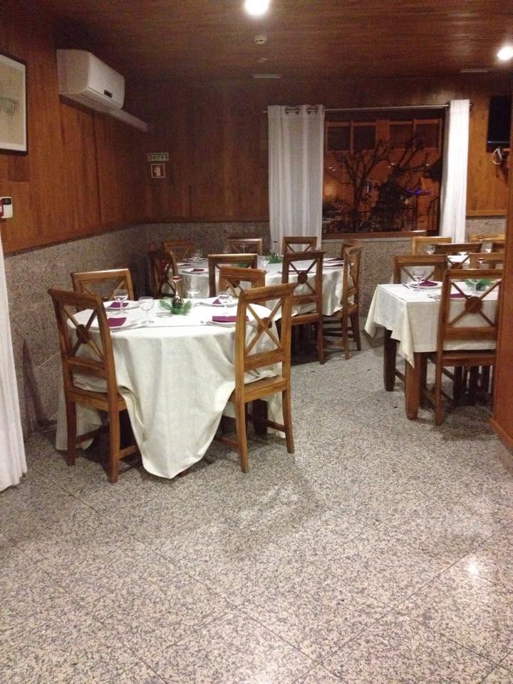 Sala de restaurante com mesas redondas cobertas por toalhas brancas, cadeiras de madeira e janela com cortinas claras.