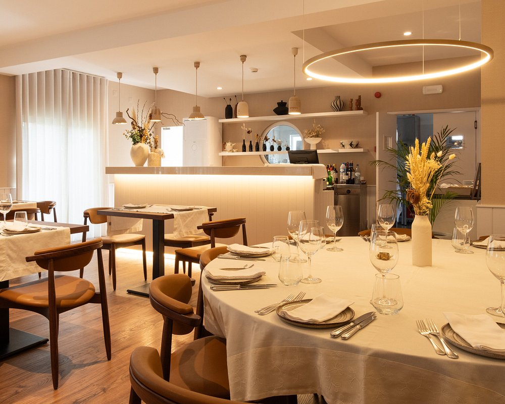 Sala de restaurante com mesas redondas e retangulares cobertas por toalhas brancas, copos, talheres, cadeiras de madeira com assentos em pele, candeeiro circular suspenso e balcão iluminado ao fundo.