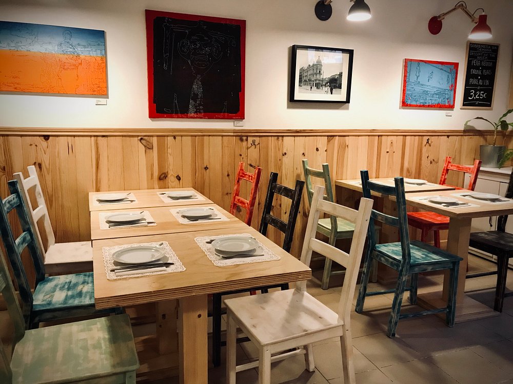 Sala com mesas de madeira, cadeiras pintadas em várias cores, pratos e talheres dispostos; paredes com quadros coloridos e fotografia a preto e branco; iluminação quente e quadro de ardósia com preço.