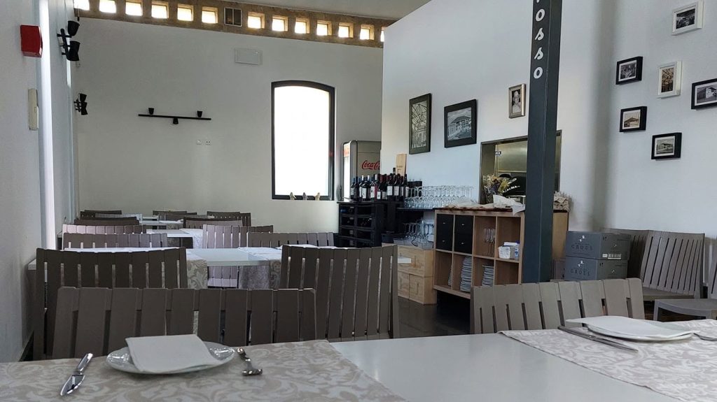 Sala com mesas de toalha clara, cadeiras de madeira, prateleiras com copos e garrafas, e frigorífico de bebidas.