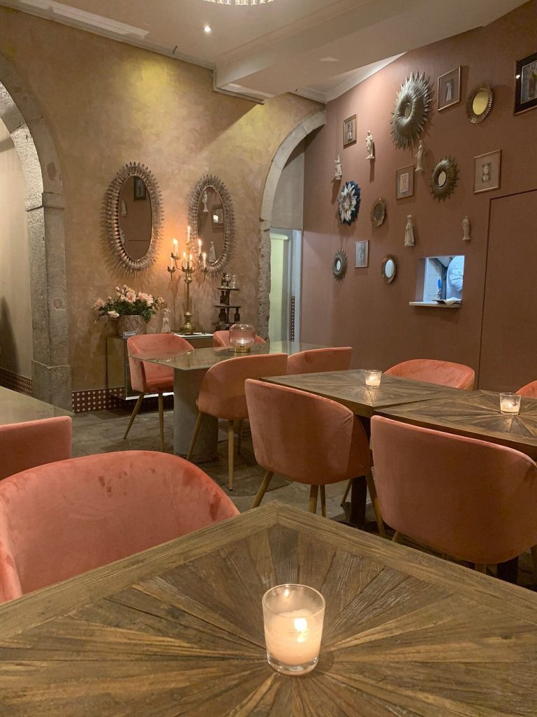 Sala com mesas de madeira, velas acesas, cadeiras rosa‑veludo, paredes em tons terra com espelhos ornamentados, quadros e esculturas; candelabro e arranjo floral sobre mesa lateral; arcadas de pedra.