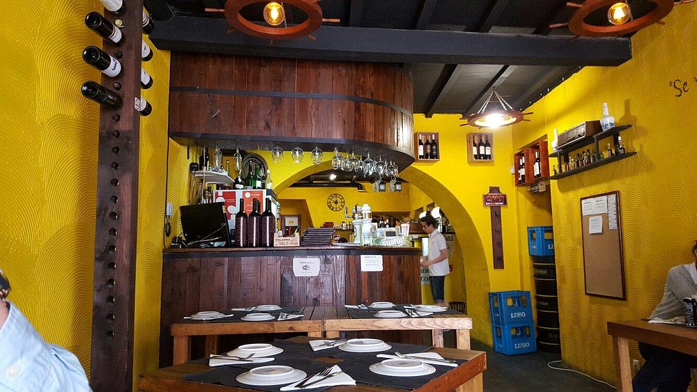 Interior da Taberna Açor com paredes amarelas, balcão em madeira, garrafeira, mesas montadas e elementos decorativos rústicos.