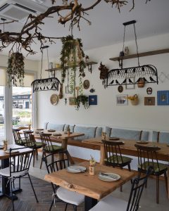 Sala com mesas de madeira, cadeiras metálicas com almofadas verdes e brancas, banco com almofadas azuis, prateleiras com cerâmica e plantas, iluminação decorativa com elementos naturais suspensos.