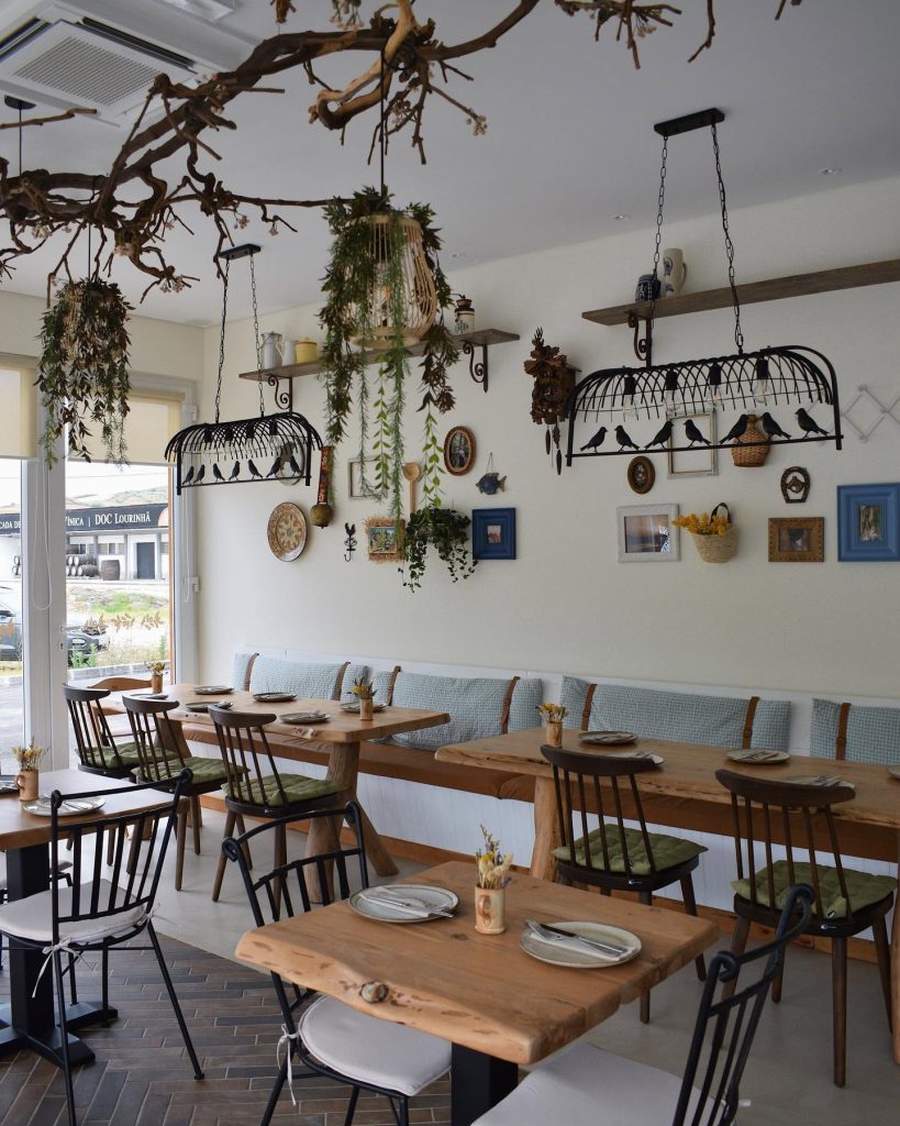 Sala com mesas de madeira, cadeiras metálicas com almofadas verdes e brancas, banco com almofadas azuis, prateleiras com cerâmica e plantas, iluminação decorativa com elementos naturais suspensos.