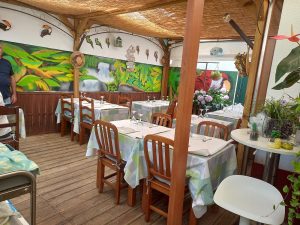 Esplanada coberta com pergola de madeira e teto entrançado; mesas com toalhas verdes e azuis, copos virados, mural tropical com aves e plantas; elementos decorativos como casinhas, plantas e placa de madeira.