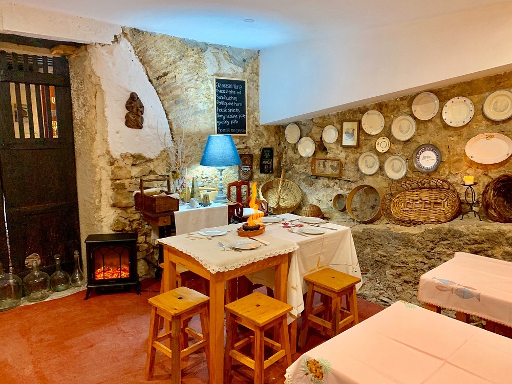 Sala com paredes de pedra, mesas com toalhas brancas, bancos de madeira, candeeiros de cúpula, lareira elétrica, prateleiras com garrafas e objetos decorativos, quadros e cestos pendurados.
