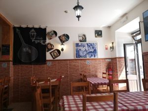 Sala com mesas de madeira e toalhas axadrezadas, paredes com fotografias, quadros e tapeçaria alusiva ao fado, iluminação quente e porta aberta para o exterior.
