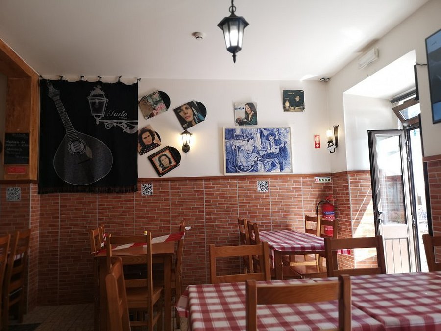 Sala com mesas de madeira e toalhas axadrezadas, paredes com fotografias, quadros e tapeçaria alusiva ao fado, iluminação quente e porta aberta para o exterior.