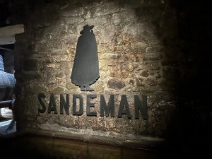Silhueta preta de figura com capa e chapéu sobre a palavra “SANDEMAN”, aplicada em parede de pedra com iluminação quente.