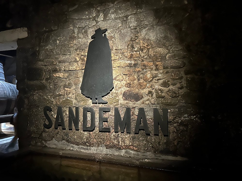 Silhueta preta de figura com capa e chapéu sobre a palavra “SANDEMAN”, aplicada em parede de pedra com iluminação quente.