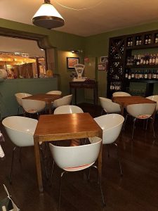 Sala com mesas de madeira, cadeiras brancas de design curvo, parede verde com garrafeira, balança antiga decorativa e iluminação pendente.