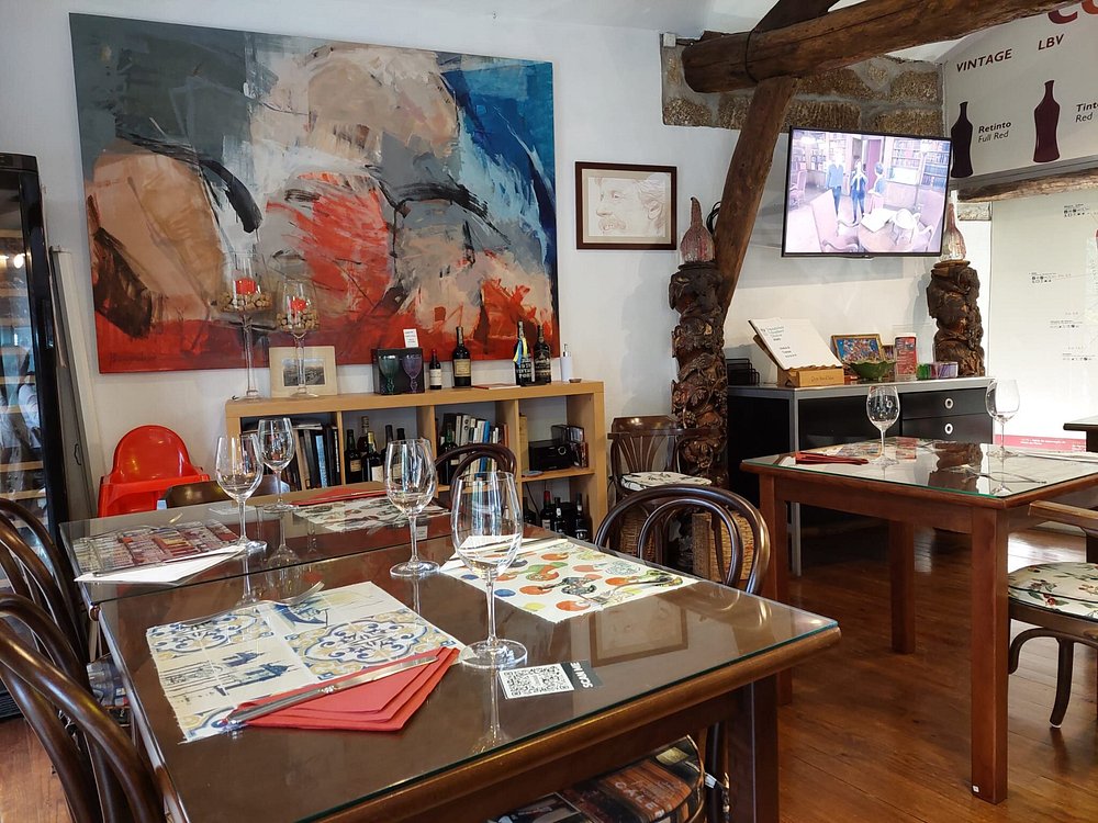 Sala com mesas de madeira, copos, guardanapos vermelhos, estante com vinhos, livros e quadro abstrato.