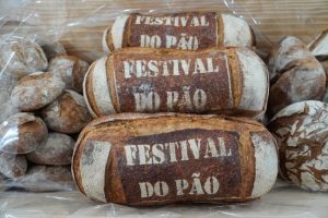Três pães grandes com a inscrição “Festival do Pão” na crosta, rodeados por pães rústicos mais pequenos.