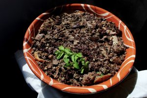 Taça de barro com prato de carne em molho escuro, decorado com ervas frescas.