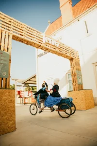 Dois artistas em trajes históricos pedalam triciclo com compartimento traseiro sob arco de madeira “CITY”; prédio com letreiro “Miranda” ao fundo.