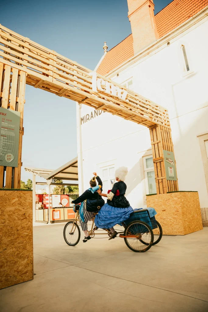 Dois artistas em trajes históricos pedalam triciclo com compartimento traseiro sob arco de madeira “CITY”; prédio com letreiro “Miranda” ao fundo.