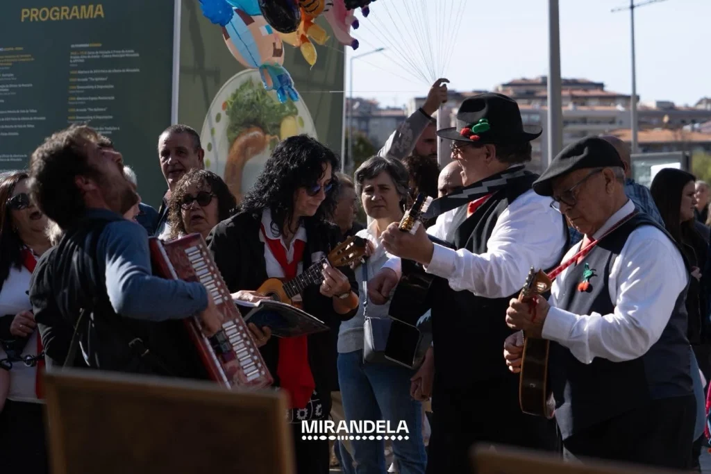Grupo de músicos com violão, acordeão e ukulele, vestidos com coletes e lenços vermelhos, tocando diante de público atento; texto “Mirandela