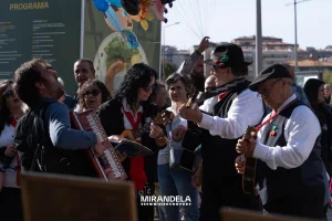 Grupo de músicos com violão, acordeão e ukulele, vestidos com coletes e lenços vermelhos, tocando diante de público atento; texto “Mirandela