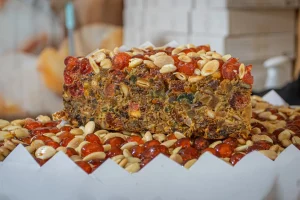 Bolo de frutas denso, coberto com cerejas cristalizadas vermelhas e nozes inteiras, em close-up com borda de papel branco.