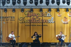 Três músicos tocam violão em palco diante de painel amarelo com textos e imagens alusivas à doçaria tradicional portuguesa.