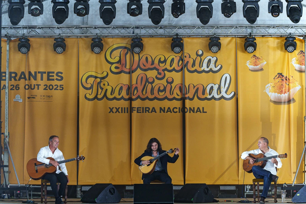 Três músicos tocam violão em palco diante de painel amarelo com textos e imagens alusivas à doçaria tradicional portuguesa.