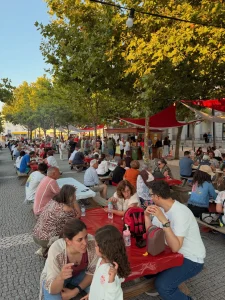 Pessoas sentadas em longas mesas ao ar livre sob árvores em praça pavimentada.