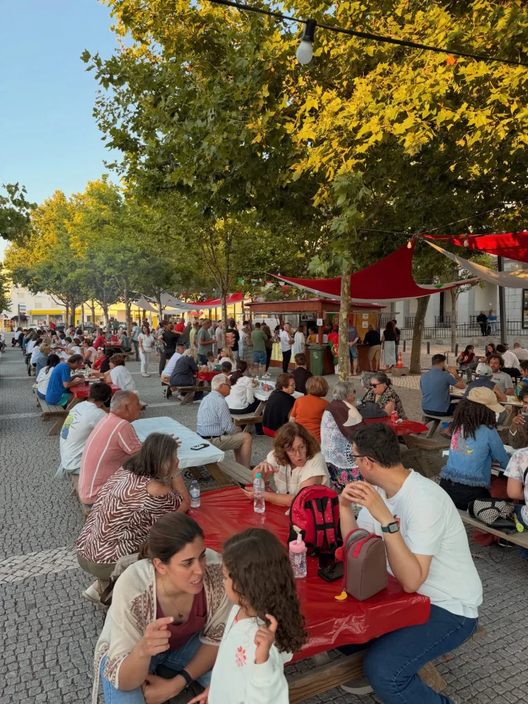 Pessoas sentadas em longas mesas ao ar livre sob árvores em praça pavimentada.