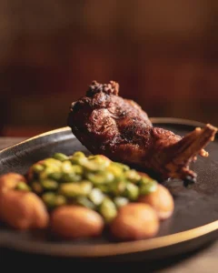 cabrito assado coberto por molho escuro, servido com batatas pequenas douradas e legumes verdes em prato escuro com borda dourada sobre mesa de madeira.