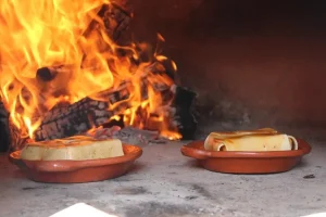 Dois recipientes de cerâmica com blocos de alimentos retangulares cobertos por calda dourada sobre a base de pedra de um forno a lenha com chamas intensas ao fundo