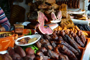 Mesa exibe presuntos defumados, bacon e variados enchidos; pratos com azeitonas e carnes fatiadas, fundo com produtos pendurados e texto “VIEIRA DO MINHO