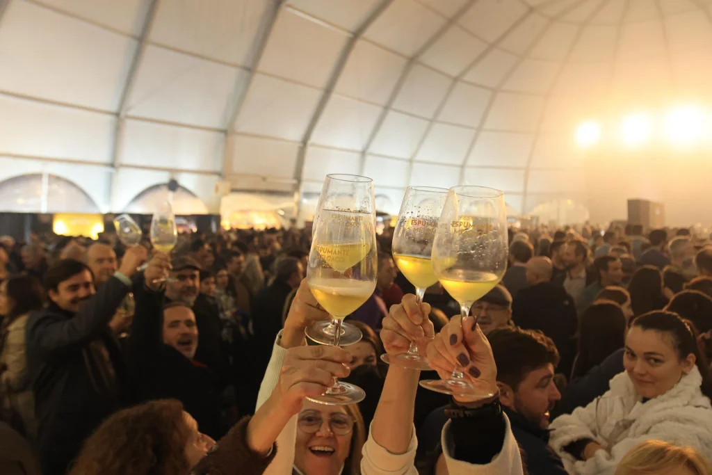 Mãos erguem taças com bebida dourada e inscrição “Festival do Espumante” em tenda cheia de pessoas celebrando.