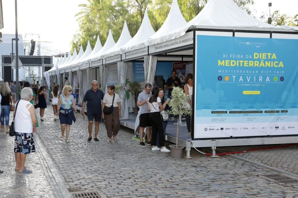 Tendas brancas alinhadas em rua de pedra, visitantes caminhando e grande banner anunciando a XI Feira da Dieta Mediterrânica em Tavira, com datas e atrações musicais.