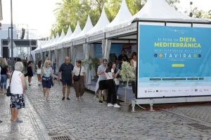 Tendas brancas alinhadas em rua de pedra, visitantes caminhando e grande banner anunciando a XI Feira da Dieta Mediterrânica em Tavira, com datas e atrações musicais.