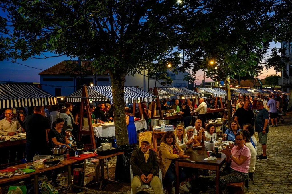Mercado noturno com barracas listradas azul e branco iluminadas; pessoas comem e conversam em mesas de madeira sob árvore central, com céu rosado ao fundo.