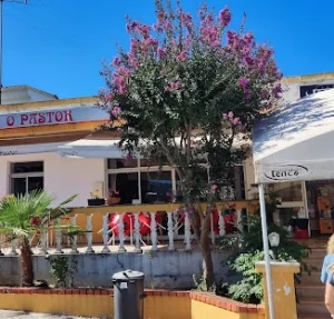 Entrada do restaurante O Pastor com letreiro vermelho, terraço com balaustrada branca e uma árvore florida com flores cor-de-rosa em destaque sob o céu azul.