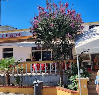Entrada do restaurante O Pastor com letreiro vermelho, terraço com balaustrada branca e uma árvore florida com flores cor-de-rosa em destaque sob o céu azul.