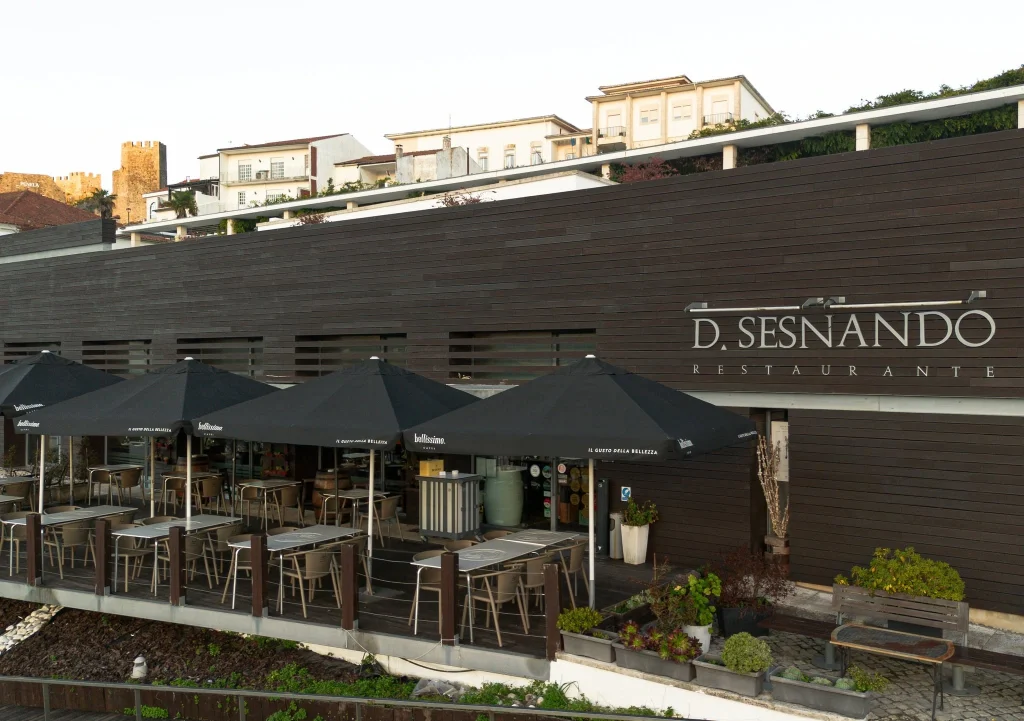 Fachada moderna em madeira do Restaurante D. Sesnando com nome em letras prateadas, esplanada externa coberta por guarda-sóis pretos e vista ao fundo para o castelo.