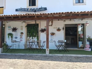 Fachada de estilo rústico da Taberna Maria Chitas, com paredes brancas, alpendre de madeira, pequenas mesas na esplanada sobre relva artificial e placas decorativas.