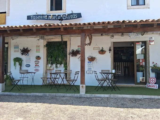 Fachada de estilo rústico da Taberna Maria Chitas, com paredes brancas, alpendre de madeira, pequenas mesas na esplanada sobre relva artificial e placas decorativas.