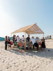 Grupo de pessoas reunido sob uma estrutura com teto de palha na areia da praia durante evento de degustação.