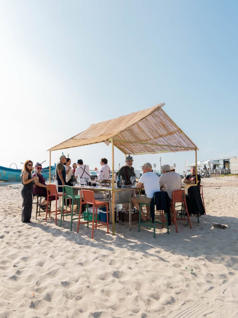 Grupo de pessoas reunido sob uma estrutura com teto de palha na areia da praia durante evento de degustação.