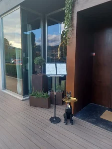 Fachada envidraçada com reflexo de árvores, vasos com plantas, suporte de menu e estátua preta de cão egípcio com bandeja dourada.