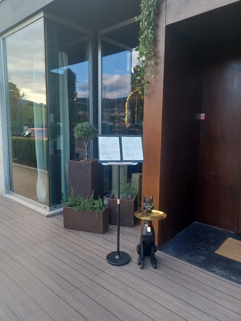 Fachada envidraçada com reflexo de árvores, vasos com plantas, suporte de menu e estátua preta de cão egípcio com bandeja dourada.
