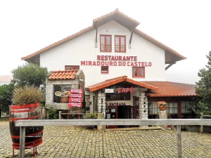 Prédio branco com telhado vermelho, letreiro “Restaurante Miradouro do Castelo”, entrada com placas “Café” e “Aberto”, menu exposto e pátio de pedra.