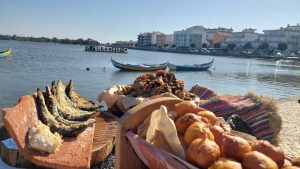 Pratos de frituras como peixes e bolinhos sobre superfícies rústicas junto ao rio, barcos portugueses ao fundo e placa “25 ANOS em celebração.