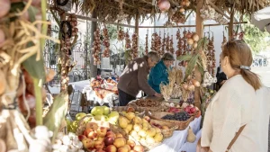 Feirantes atendem cliente em uma banca coberta de palha repleta de cestas de frutas e réstias penduradas de alho e cebola em uma feira ao ar livre.