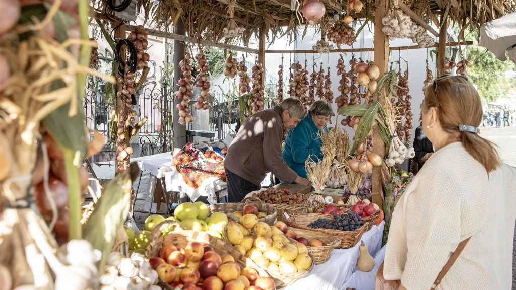 Feirantes atendem cliente em uma banca coberta de palha repleta de cestas de frutas e réstias penduradas de alho e cebola em uma feira ao ar livre.