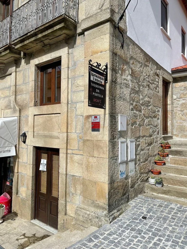 Fachada de pedra com letreiro “Restauração e Bebidas Tasquinha da Portela Bicafé”, entrada com plantas e varanda com grade metálica.
