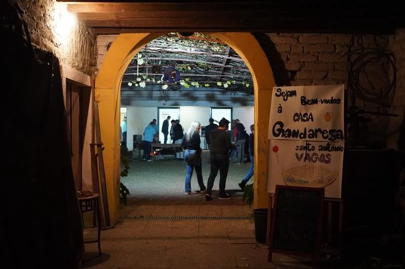 Pessoas reunidas à noite em pátio iluminado, sob arco amarelo, com teto decorado por vinhas e cartaz escrito “Sejam Bem-vindos à CASA Gandarega santo antonio VAGAS”.