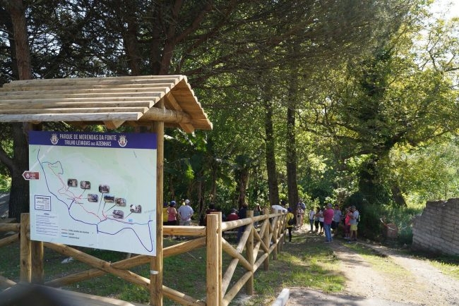 Placa coberta exibe mapa do “Parque de Merendas da Ponte Trilho Caminhos das Azenhas”, cercada por árvores e visitantes em trilha.
